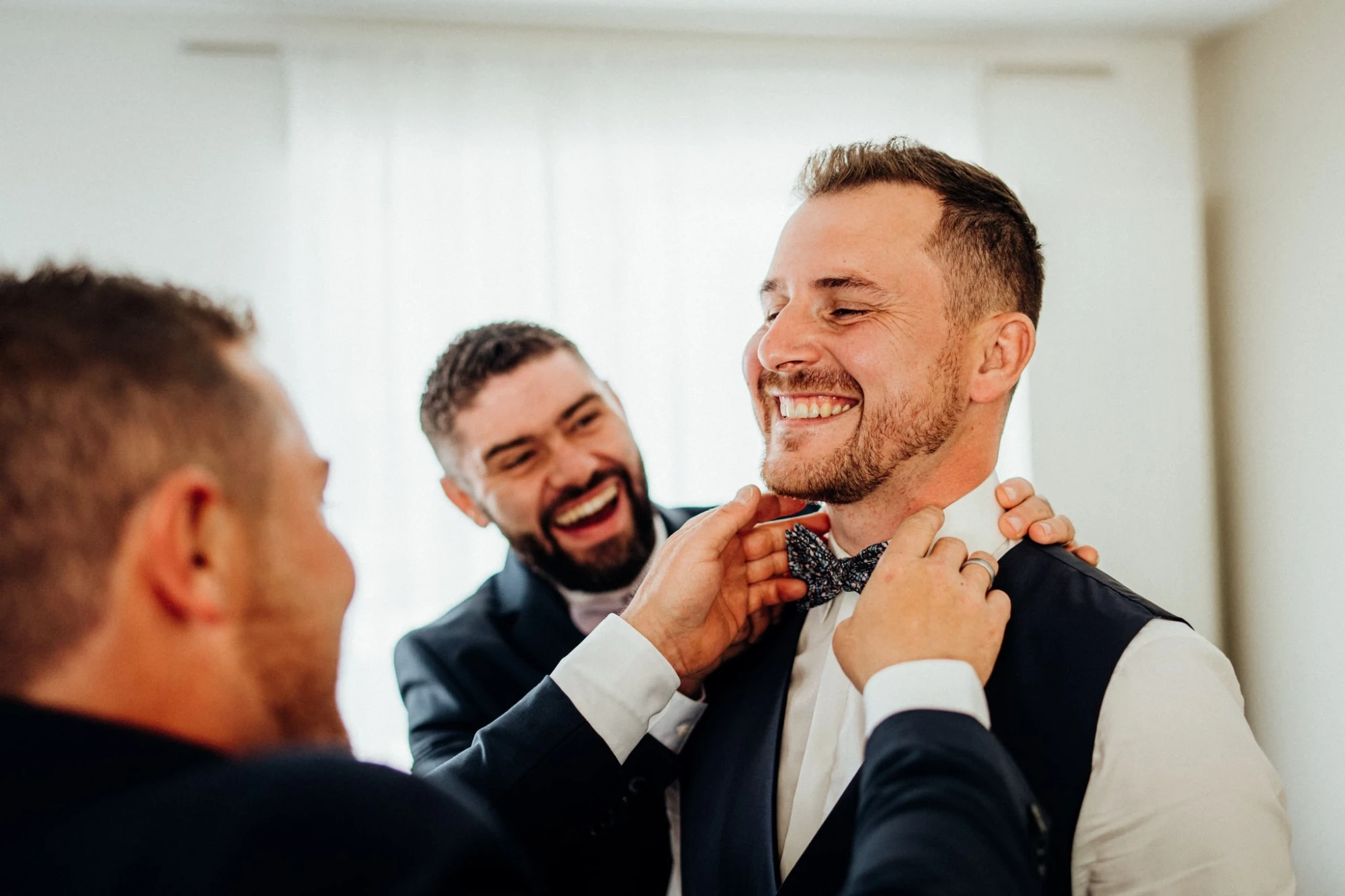 Un marié souriant se fait ajuster son nœud papillon par un ami avant le mariage. photographe-lille