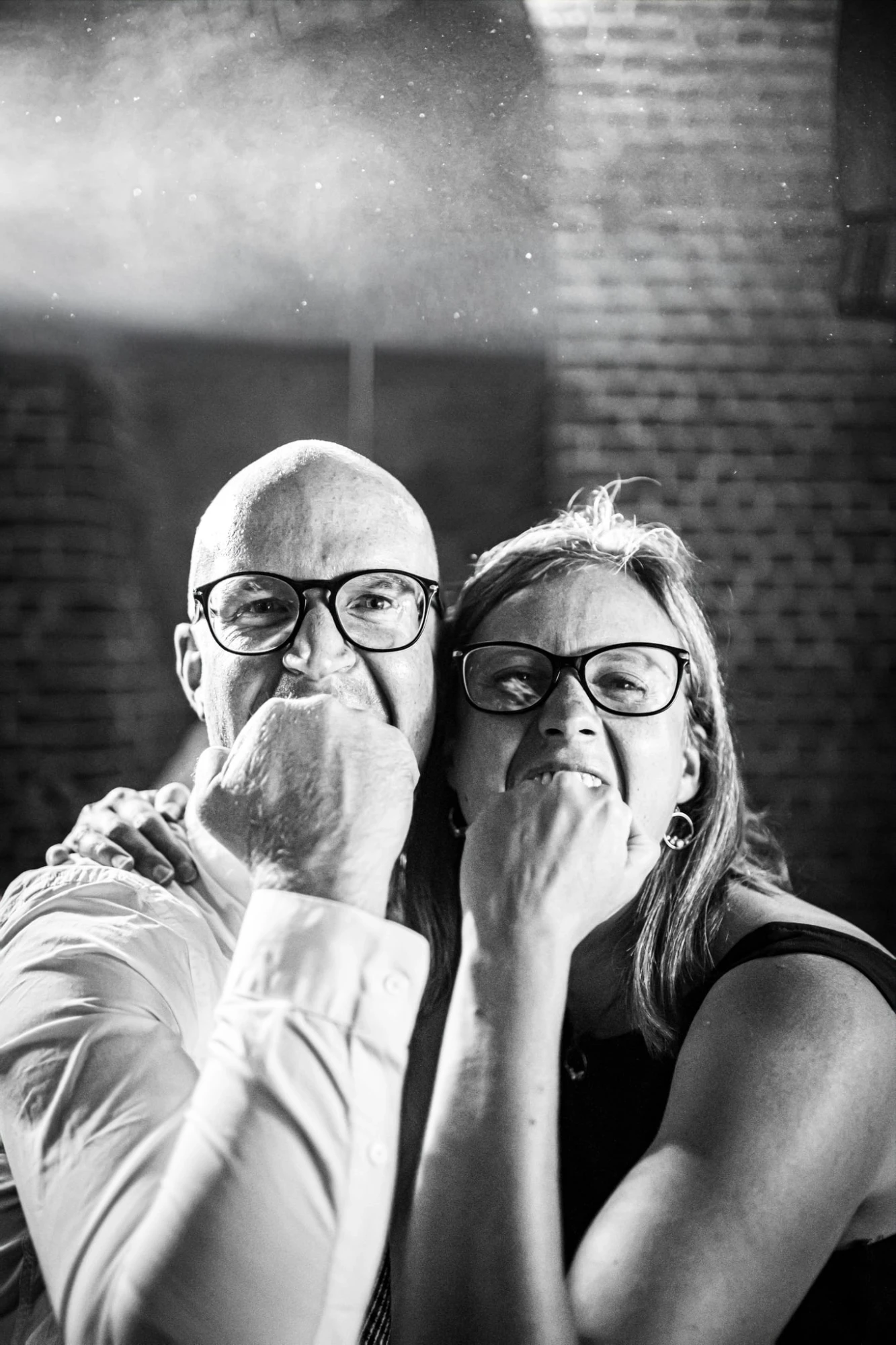 Couple souriant avec des lunettes, posant avec les mains sur le menton devant un mur de briques. photographe-lille