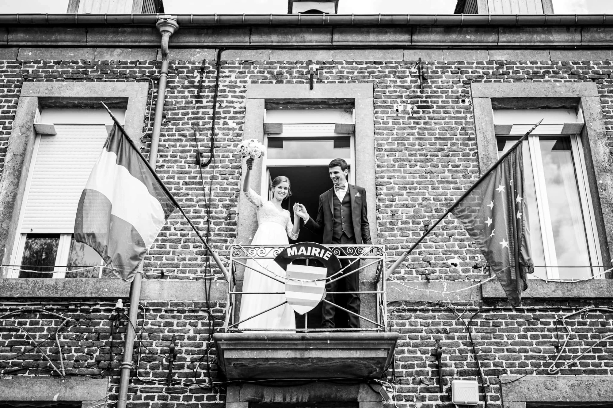 Jeunes mariés sur le balcon de la mairie avec drapeaux français et européen. "MAIRIE" visible. photographe-lille