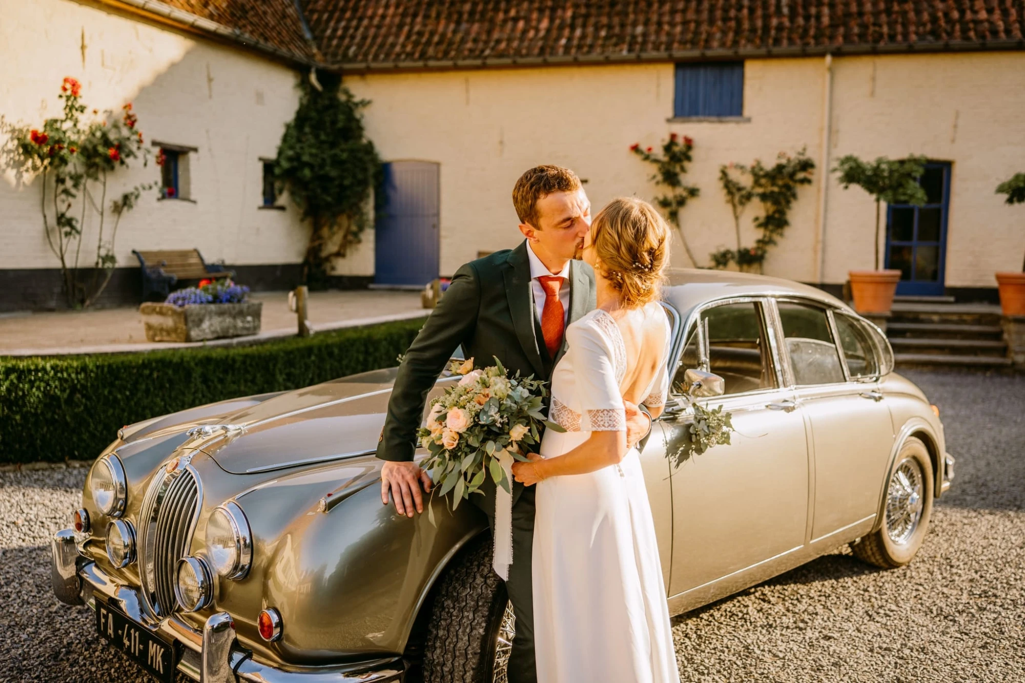 Mariés s'embrassant près d'une Jaguar classique argentée devant une ferme. photographe-lille