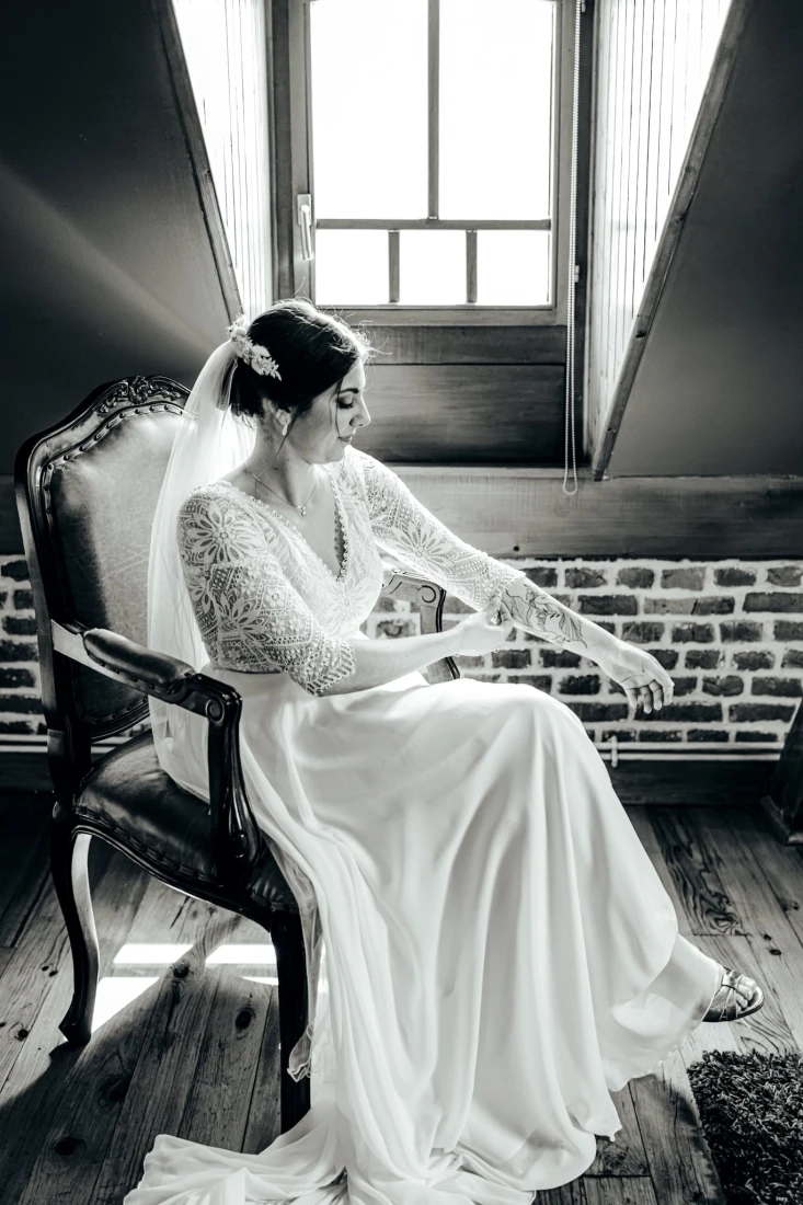 Mariée en robe blanche assise sur une chaise vintage devant une fenêtre. Détails dentelle et voile. photographe-lille