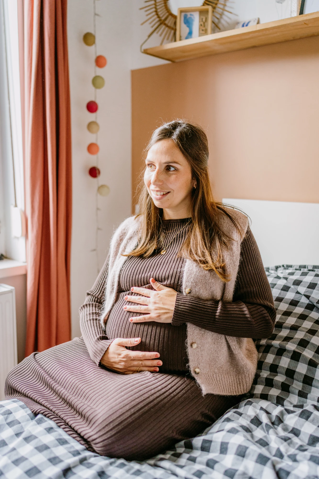 Femme enceinte souriante assise sur un lit, tenant son ventre. Maternité épanouie. photographe-lille